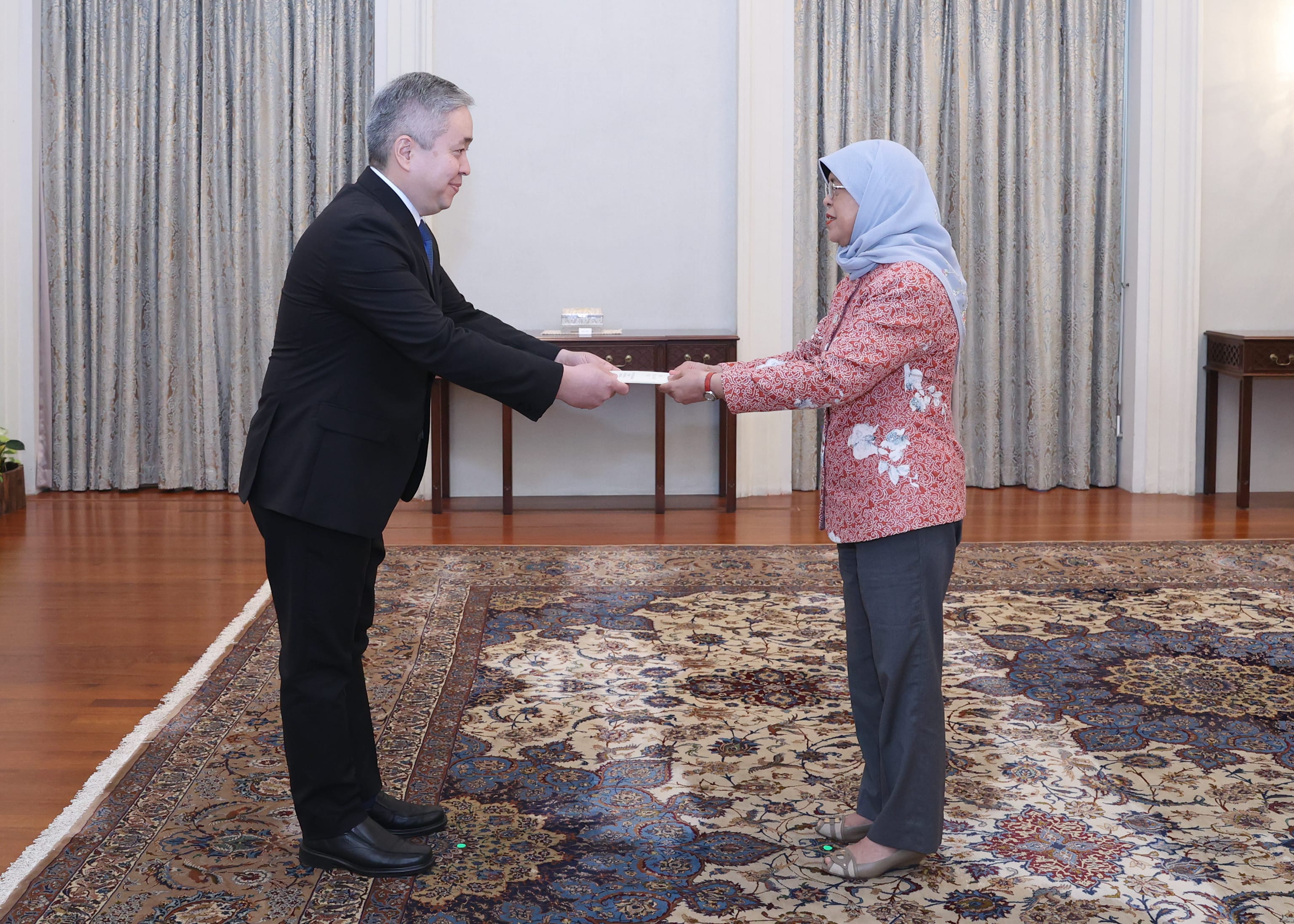Two people exchange a document; man in suit, woman in hijab.